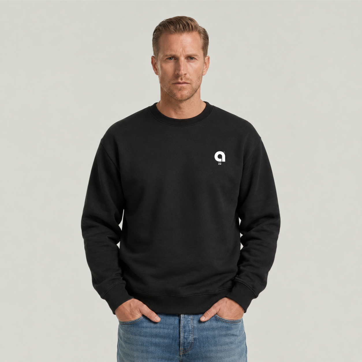 A man wearing a black crewneck sweatshirt with a small white circular logo on the left chest, paired with blue jeans, standing against a light gray studio background.