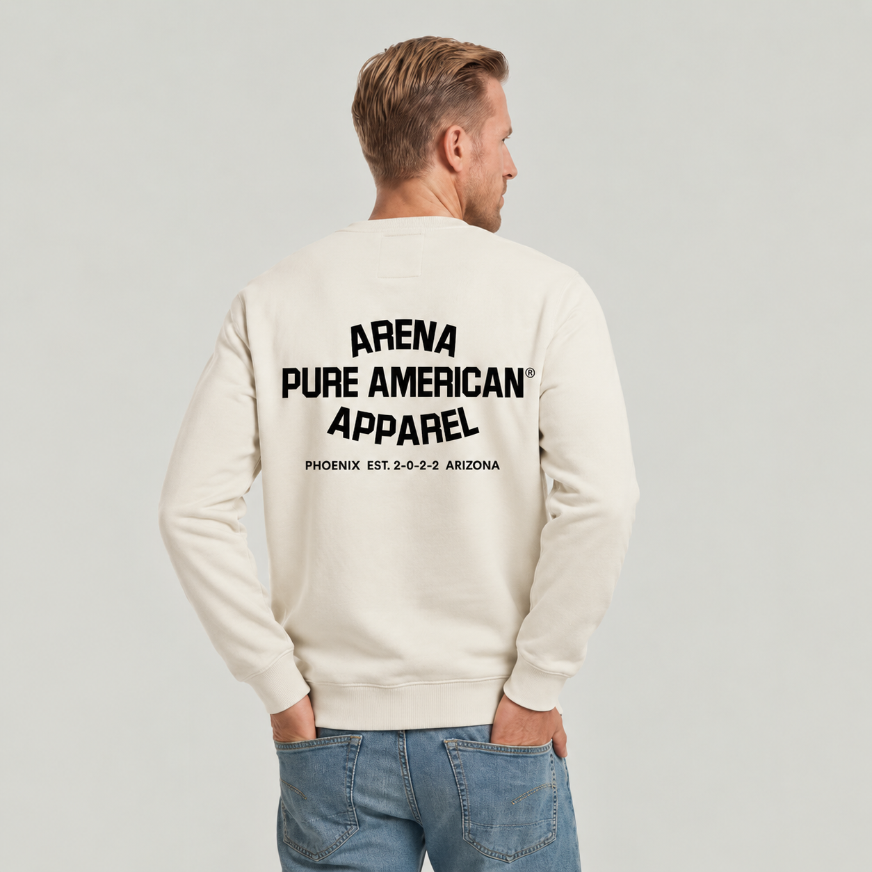 A man viewed from behind wearing an off-white crewneck sweatshirt with black text on the back reading 'ARENA PURE AMERICAN APPAREL PHOENIX EST. 2-0-2-0 ARIZONA', paired with blue jeans, against a light gray studio background.
