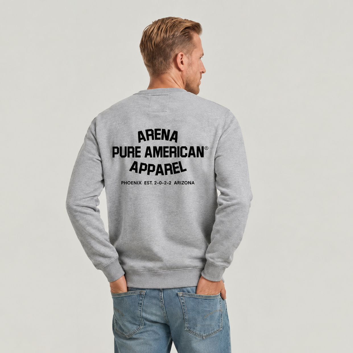 A man viewed from behind wearing a light gray crewneck sweatshirt with 'ARENA PURE AMERICAN APPAREL' printed in black on the back, paired with blue jeans, against a light gray studio background.
