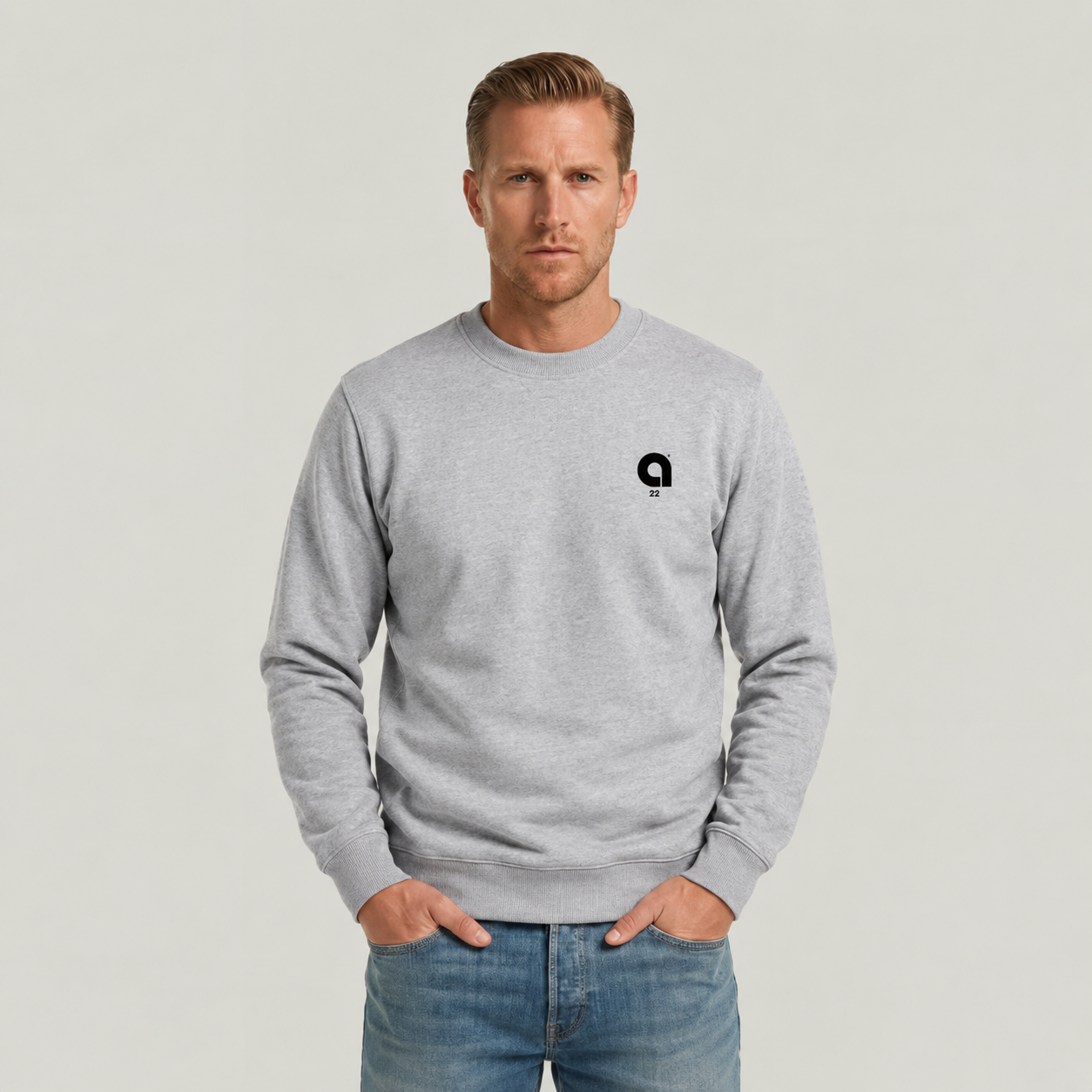 A man wearing a light gray crewneck sweatshirt with a small black circular logo on the left chest, paired with blue jeans, standing against a light gray studio background.