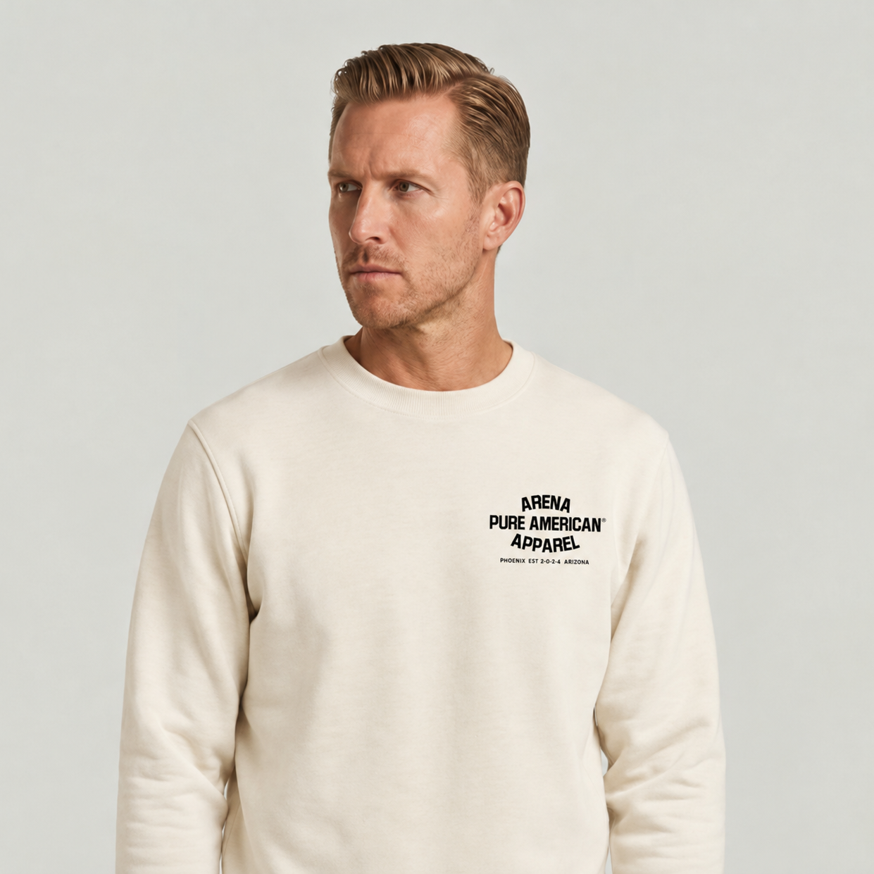 A man wearing a cream-colored crewneck sweatshirt with black text on the left chest that reads 'ARENA PURE AMERICAN APPAREL'.