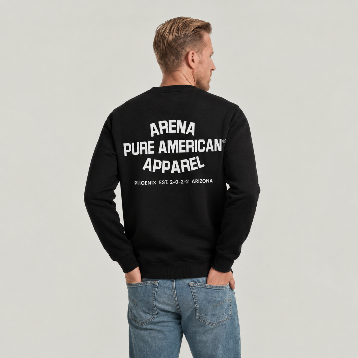 A man viewed from behind wearing a black crewneck sweatshirt with bold white text reading 'ARENA PURE AMERICAN APPAREL' and 'PHOENIX EST. 2-0-2-2 ARIZONA' printed on the back, paired with blue jeans against a light gray studio background.
